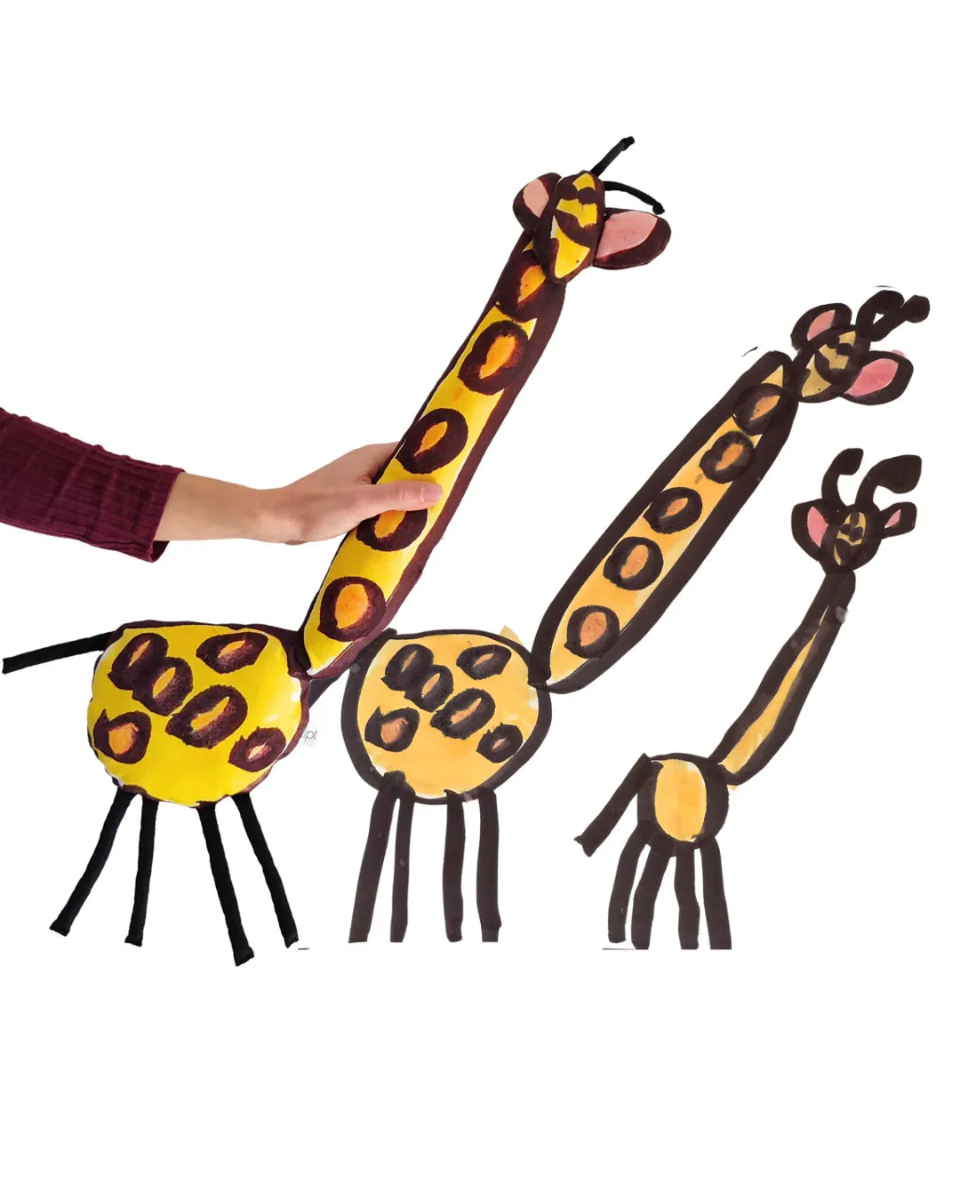 il disegno di un bambino raffigurante una giraffa è stato cucito a mano e trasformato fedelmente in un pupazzo di nome monamì. nell'immagine le due versioni a confronto: il disegno originale realizzato con pennarelli su carta e il pupazzo stampato digitalmente su tessuto twill di cotone e cucito a mano. le giraffe hanno un collo monto lungo e grandi orecchia rosa a disco. il corpo si regge su gracili zampe nere.