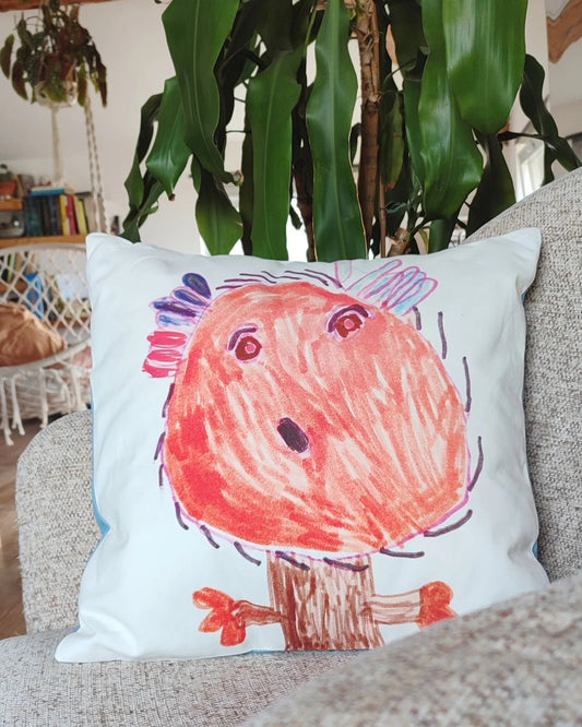 il disegno di un personaggio di fantasia realizzato da un bambino con i pennarelli colorati stampato in digitale su di un cuscino 40x40 in cotone certificato. il cuscino è su una poltrona all'interno di un salotto come complemento d'arredo.